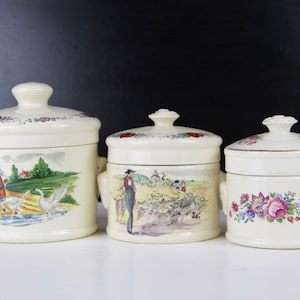 May include: Three white ceramic canisters with lids, each featuring a different floral or countryside scene. The largest canister has a scene of a woman and two geese in a field, the middle canister has a scene of a man and a flock of geese, and the smallest canister has a floral design with pink roses.