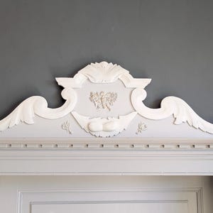 May include: A white decorative architectural molding with ornate scrollwork and a central design. The molding is mounted above a door frame, with a gray wall in the background. The design includes floral and leaf motifs.