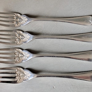 Può includere: Quattro forchette d'argento con dettagli floreali e volute ornamentali sui manici. I rebbi sono lunghi e sottili, e i manici hanno un design a forma di cuore all'estremità. Le forchette sono disposte in fila su una superficie grigia.