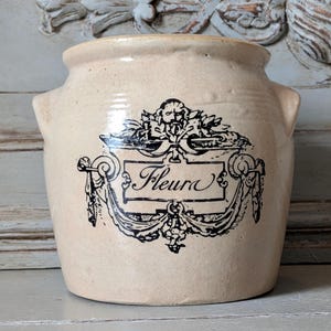 Puede incluir: Una vasija de cerámica beige con dos asas laterales y un gráfico impreso en negro en la parte frontal. El diseño presenta un marco ornamentado con un motivo central de cabeza de león y la palabra Fleura escrita en cursiva en un cartel.