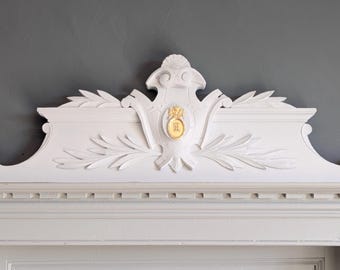 Può includere: Dettaglio architettonico bianco e ornato con stemma centrale e decorazioni a foglie. Lo stemma presenta un emblema dorato con un'iniziale. Il design è posto su uno sfondo grigio, evidenziando le intricate sculture.