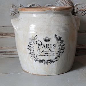 Peut inclure: Un pot en céramique de couleur crème avec deux anses latérales et un large rebord. L'avant présente un motif imprimé noir représentant une couronne au-dessus du texte Paris Exposition Universelle, entouré d'une couronne de feuilles et branches.