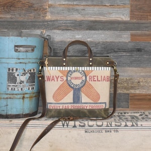 May include: A tote bag crafted from tan canvas and dark olive green leather, featuring a vintage-style graphic of two corn cobs and the text "FARMCRAFT HYBRIDS OXFORD IND ALWAYS RELIABLE EVERY EAR PROPERLY CROSSED" with brown leather handles and strap.