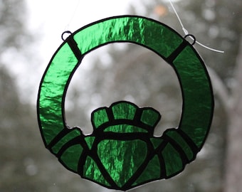 May include: A green stained glass Celtic knot, also known as a Claddagh, in the shape of a heart with two hands holding it. The knot is a traditional Irish symbol of love, friendship, and loyalty.