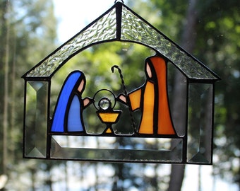 May include: Stained glass nativity scene with a house-shaped frame. The figures are depicted in blue and orange, with a central figure in a gold-colored cradle. The frame is black with clear glass panels.