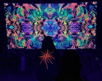 May include: A person wearing a black hoodie with a glowing orange starburst design stands in front of a large, colorful tapestry with psychedelic patterns. The tapestry is lit with blacklight, making the colors glow brightly.