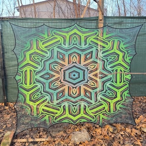 May include: A large, colorful mandala-style tapestry with a green, orange, and blue color scheme. The tapestry is made of a thin, mesh-like fabric and is hanging from a fence.