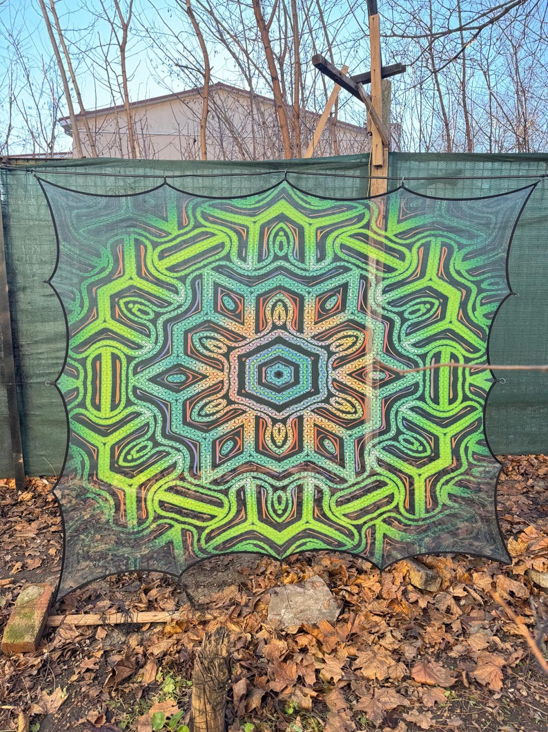 May include: A large, colorful mandala-style tapestry with a green, orange, and blue color scheme. The tapestry is made of a thin, mesh-like fabric and is hanging from a fence.