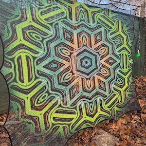 May include: A black mesh tapestry with a colorful geometric design. The design features a central mandala with green, orange, and blue colors. The tapestry is hanging outdoors.