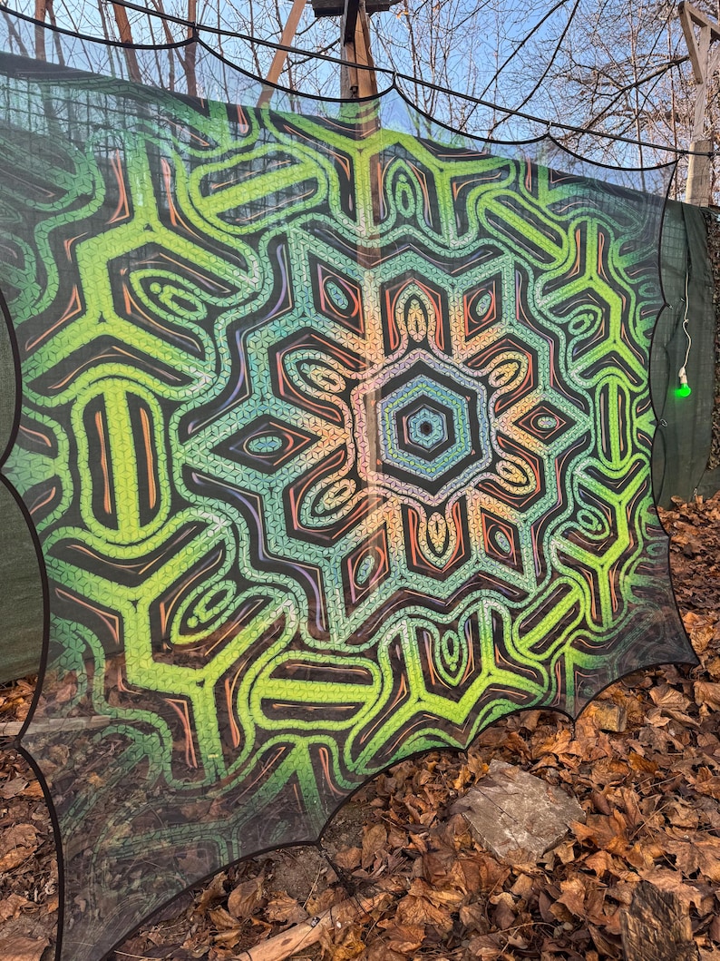 May include: A black mesh tapestry with a colorful geometric design. The design features a central mandala with green, orange, and blue colors. The tapestry is hanging outdoors.