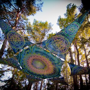 May include: A colorful tapestry with geometric patterns hangs between trees in a forest. The tapestry is blue, green, yellow, and pink.