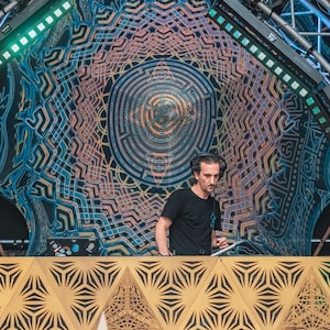 May include: A DJ performs at a music festival, standing behind a wooden DJ booth with a geometric design. The DJ is wearing a black shirt and headphones. The background features a large, colorful tapestry with a geometric pattern.