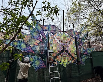 May include: A colorful, psychedelic tapestry is being hung on a fence. The tapestry is made of several triangular pieces, each with a different design. The tapestry is being hung by two people, one on a ladder and one on the ground.
