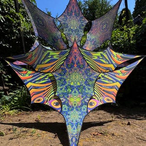 Puede incluir: Un gran toldo geométrico y colorido con un diseño en forma de estrella. El toldo presenta patrones vibrantes en tonos de azul, verde, naranja y morado. El diseño incluye formas de ojo y detalles intrincados al estilo mandala, creando un efecto psicodélico.