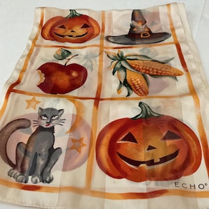 Peut inclure: Un foulard crème avec un motif quadrillé présentant des illustrations sur le thème d'Halloween. Le foulard comprend des images de citrouilles, un chapeau de sorcière, une pomme, du maïs, un chat noir et des étoiles. La marque "ECHO" est imprimée en bas à droite.
