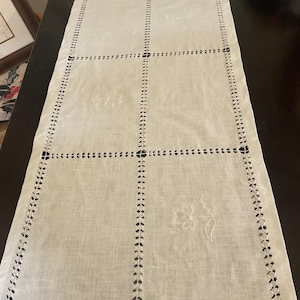 May include: A white rectangular table runner with a grid pattern and embroidered floral details. The edges and grid lines feature a cut-out design. The runner is made of a light-colored fabric and is placed on a dark surface.