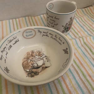 May include: A white ceramic bowl and matching mug featuring Peter Rabbit illustrations. The bowl displays three rabbits, a basket, and text reading Peter Rabbit and They went down the lane to gather blackberries. The mug features similar text.