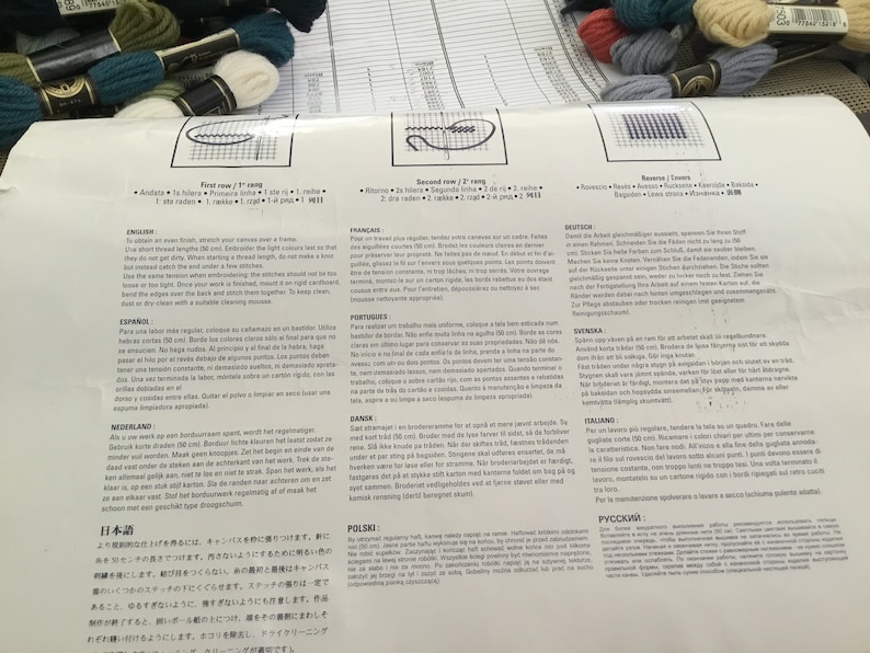 May include: A multi-lingual instruction sheet for a cross-stitch embroidery kit. The sheet includes instructions in English, French, German, Dutch, Spanish, Portuguese, Italian, Polish, and Japanese. The sheet also includes a diagram of the cross-stitch pattern.