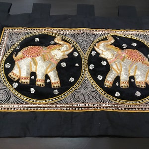 May include: A black velvet tapestry with two embroidered white elephants in gold and silver circles. The elephants are decorated with pink, red, and gold sequins.