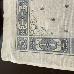May include: Close-up of a white handkerchief with a detailed border design in shades of gray. The border features floral and geometric patterns, with a delicate, ornamental corner flourish. The fabric has a slightly textured appearance.