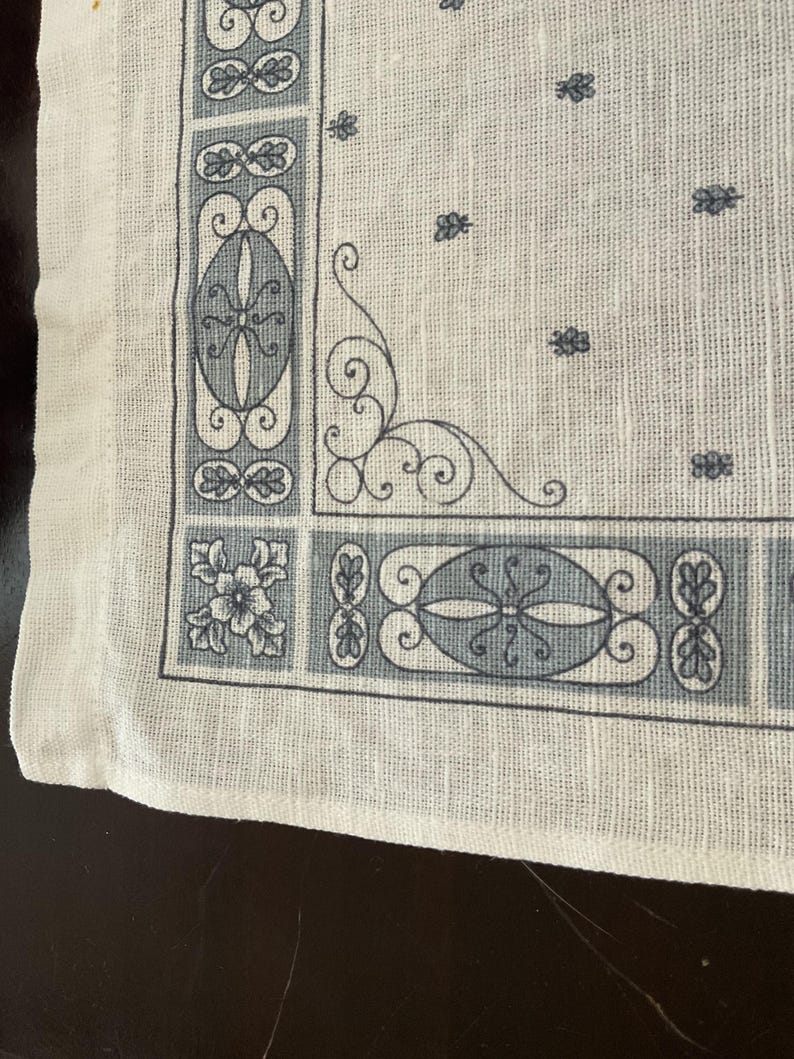 May include: Close-up of a white handkerchief with a detailed border design in shades of gray. The border features floral and geometric patterns, with a delicate, ornamental corner flourish. The fabric has a slightly textured appearance.