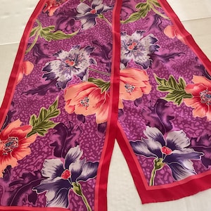 May include: A long, rectangular scarf with a vibrant floral pattern. The scarf features large flowers in shades of pink, purple, and peach, with green leaves against a magenta background. The edges are trimmed with a solid red border.