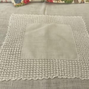 May include: A white square handkerchief with a crocheted border. The center of the handkerchief is plain, while the edges feature a delicate, openwork design. The handkerchief is made of a soft, lightweight fabric.