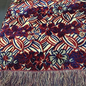 May include: A floral patterned throw blanket with a cream background, red and blue flowers, and blue leaves. The blanket has a fringe border with cream, red, and blue threads. The blanket is folded.