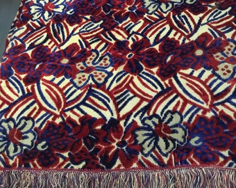 May include: A floral patterned throw blanket with a cream background, red and blue flowers, and blue leaves. The blanket has a fringe border with cream, red, and blue threads. The blanket is folded.