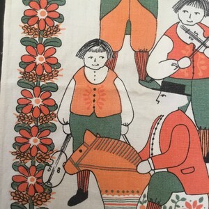 May include: A vintage fabric panel with a folk art design featuring four figures. Two figures are playing musical instruments, one figure is riding a rocking horse, and one figure is standing with a musical instrument. The background is a white fabric with a border of red flowers.
