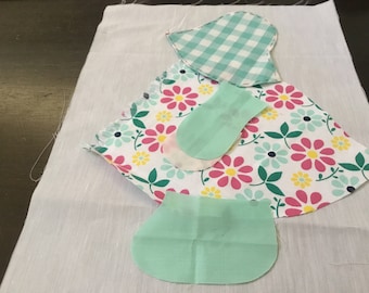 May include: Fabric pieces for a doll dress, including a floral print skirt, a gingham hat, and mint green shoe-shaped pieces. The fabric is laid out on a white background.