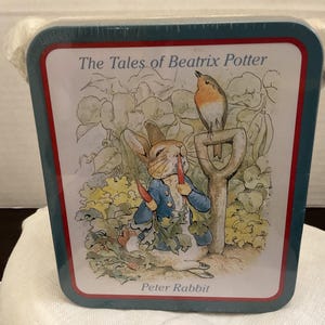 May include: A rectangular metal tin featuring a Peter Rabbit illustration from The Tales of Beatrix Potter. The image shows a rabbit in a blue jacket eating a carrot next to a garden shovel and a bird. The tin has a dark teal border and red trim.