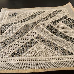 Könnte beinhalten: Eine detaillierte Gobelin-Leinwand mit einem geometrischen Design in Schwarz, Beige und Weiß. Das Muster umfasst verschiedene Stichtechniken, die ein strukturiertes Aussehen erzeugen. Ein kleiner Schmetterling ist in der oberen linken Ecke gestickt.