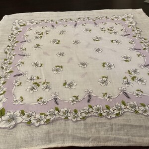May include: A square white handkerchief featuring a lavender border with a scalloped edge. The fabric is decorated with a repeating pattern of white flowers with green leaves and small dark sprigs, centered within the lavender frame.