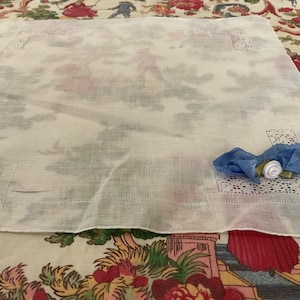 May include: A white linen handkerchief with a delicate, embroidered border and a faint, faded pattern. A blue ribbon and a small white and green floral embellishment are placed on the corner. The handkerchief is displayed on a floral patterned surface.