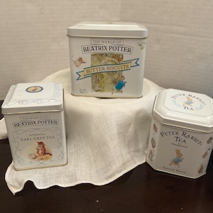 May include: Three white metal tins featuring Beatrix Potter illustrations. The center tin is labeled The World of Beatrix Potter Butter Biscuits. The left tin is labeled The World of Beatrix Potter Authentic Earl Grey Tea. The right tin is Peter Rabbit Tea.