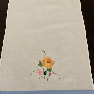 May include: White linen towel with an embroidered floral design. The embroidery features a large orange and yellow flower with a pink flower and green leaves. A light blue border is at the bottom.