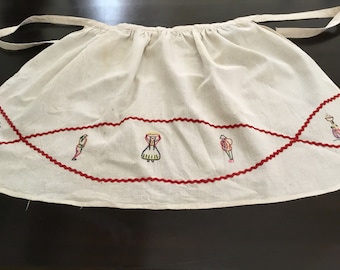 May include: A white linen apron with red embroidered trim and a design of four figures in colorful clothing.