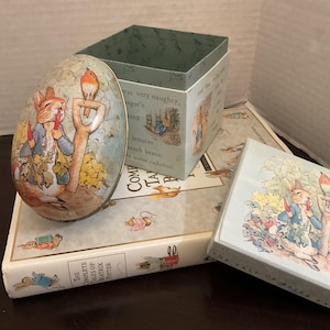 May include: An egg-shaped tin, a square box, and a book featuring Peter Rabbit illustrations. The egg and box show Peter Rabbit with a shovel and bird. The book is titled The Complete Tales of Beatrix Potter and includes various character illustrations.