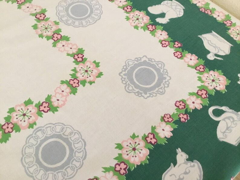 May include: A white tablecloth with a green and white floral pattern. The pattern features pink flowers, green leaves, and white teapots and cups.