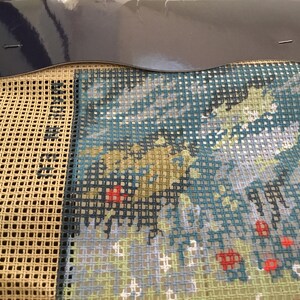 May include: A close-up of a partially completed cross-stitch project. The design features a blue, green, and white landscape with red accents. The canvas is beige and the threads are visible.