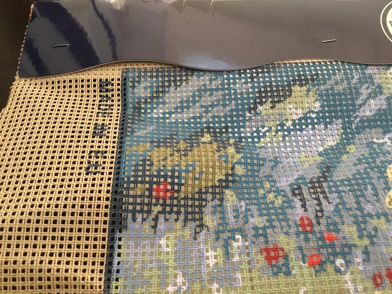 May include: A close-up of a partially completed cross-stitch project. The design features a blue, green, and white landscape with red accents. The canvas is beige and the threads are visible.