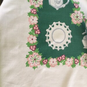 May include: A white tablecloth with a green border featuring a floral pattern and a white teacup and saucer. The floral pattern includes pink and white flowers with green leaves.