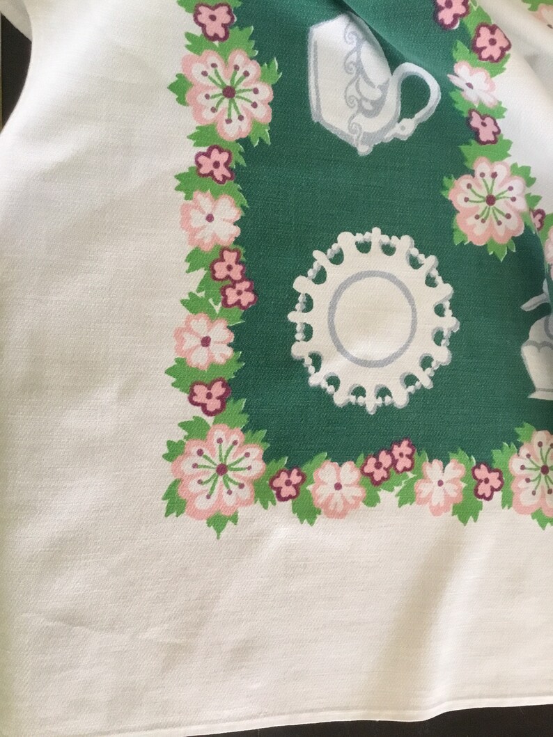 May include: A white tablecloth with a green border featuring a floral pattern and a white teacup and saucer. The floral pattern includes pink and white flowers with green leaves.