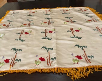 May include: A white tablecloth with a yellow fringe border features an embroidered design of stylized bamboo trees, birds, and flowers. The embroidery is done in shades of green, red, and yellow.