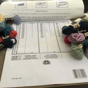 May include: A close-up of a DMC embroidery floss color chart with various skeins of floss in different colors, including red, blue, green, and brown. The chart includes the DMC floss numbers and the names of the different types of floss.