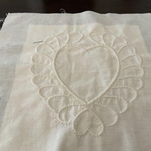 Puede incluir: Un cuadrado de tela blanca con un diseño de corazón blanco bordado en el centro. El corazón está rodeado por un borde decorativo de puntadas blancas.