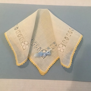 Op de afbeelding: Een wit zakdoekje met een gele gehaakte rand en een blauwe satijnen strik. Het zakdoekje heeft een delicaat kantpatroon.