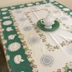 May include: A white tablecloth with a green and pink floral border. The tablecloth features a repeating pattern of teacups, saucers, and floral sprigs. Two white milk glass cream jugs are on a green plate in the centre of the tablecloth.