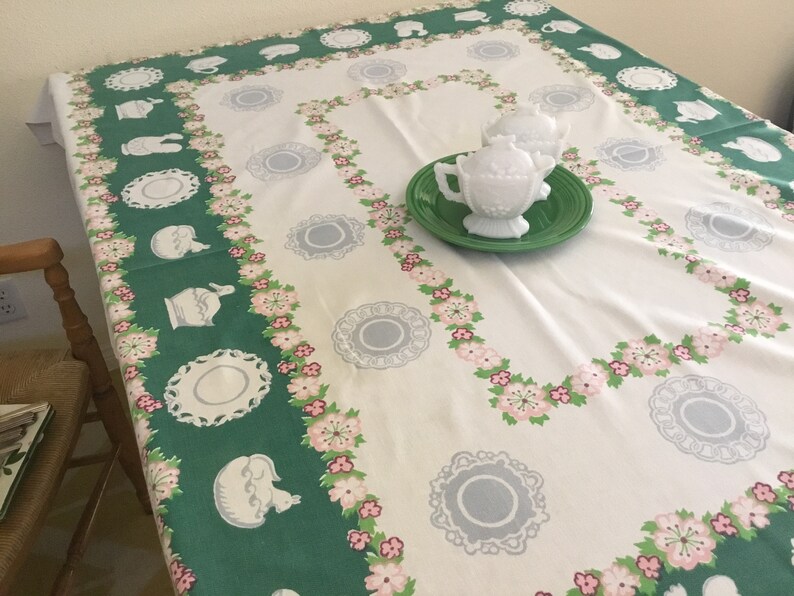 May include: A white tablecloth with a green and pink floral border. The tablecloth features a repeating pattern of teacups, saucers, and floral sprigs. Two white milk glass cream jugs are on a green plate in the centre of the tablecloth.